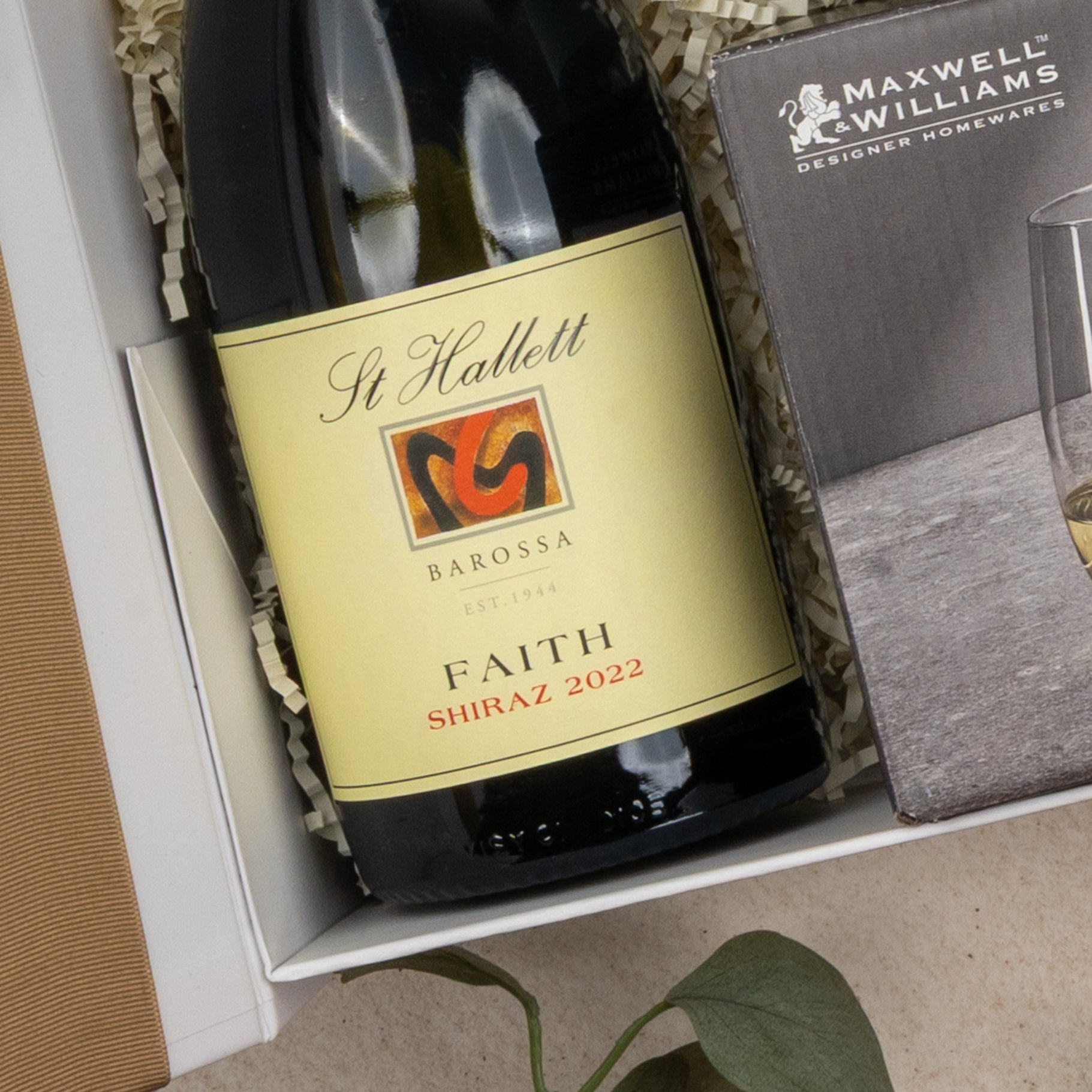The Hamper Boutique Co Relaxing Red Wine Hamper features a 2022 Shiraz wine in a gift box beside Maxwell & Williams designer glassware, with shredded paper and a green leaf for decoration.