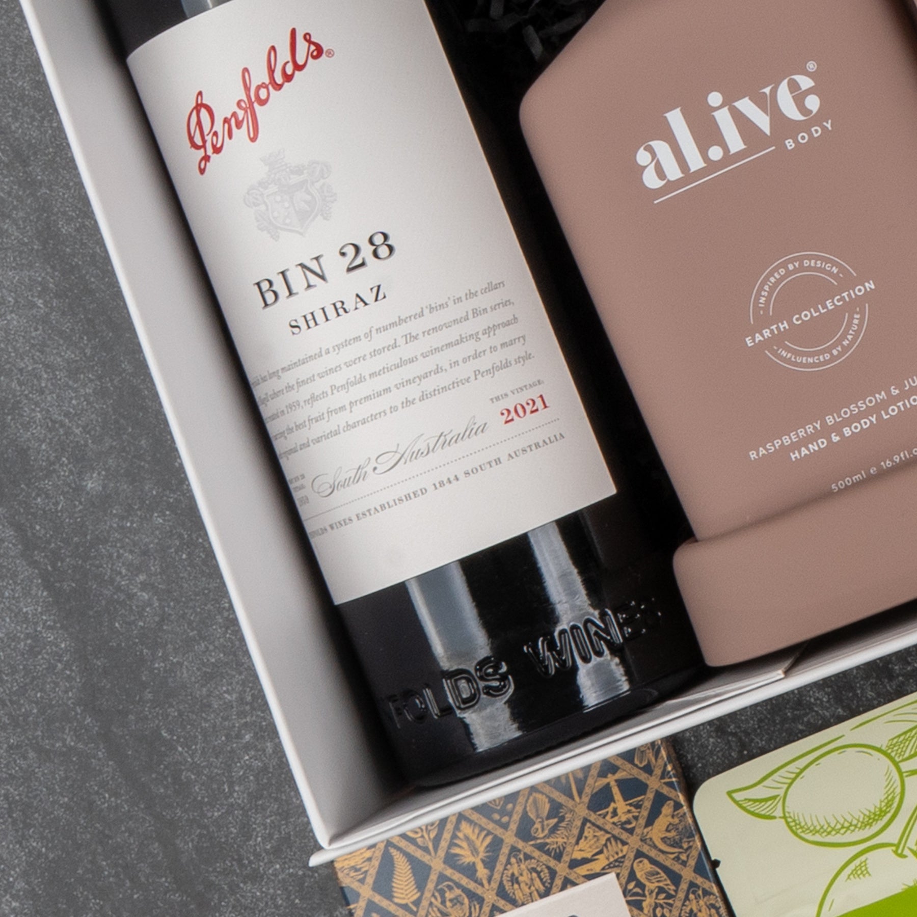 The Hamper Boutique Co Deluxe Wine and Relaxation Hamper features a bottle of Penfolds Bin 28 Shiraz red wine and a pink al.ive body lotion, artfully arranged with other luxurious gift items.