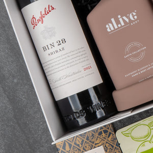 The Hamper Boutique Co Deluxe Wine and Relaxation Hamper features a bottle of Penfolds Bin 28 Shiraz red wine and a pink al.ive body lotion, artfully arranged with other luxurious gift items.