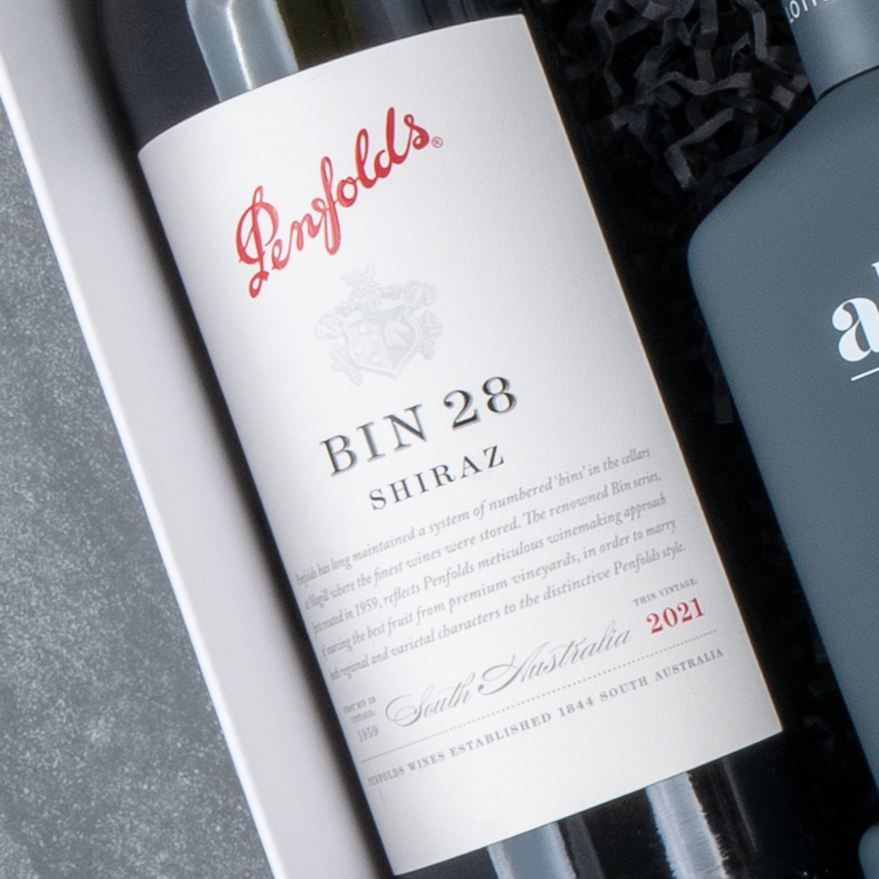 A close-up of The Hamper Boutique Co Ultimate Red Wine Celebration Hamper, featuring a 2021 Penfolds BIN 28 Shiraz bottle with a label showing red and black text, lying in a gift box beside a dark-colored wine bottle.