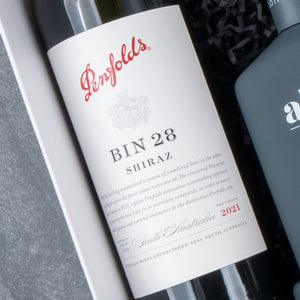 A close-up of The Hamper Boutique Co Ultimate Red Wine Celebration Hamper, featuring a 2021 Penfolds BIN 28 Shiraz bottle with a label showing red and black text, lying in a gift box beside a dark-colored wine bottle.