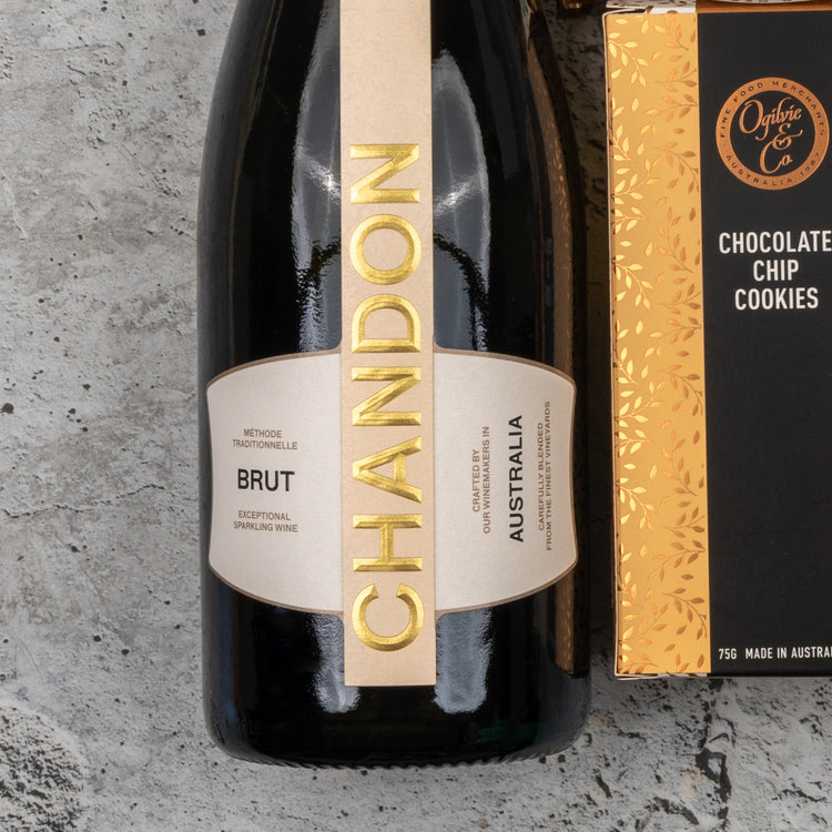A Chandon Sparkling Delights Hamper by The Hamper Boutique Co is displayed on a textured gray surface, featuring a bottle of sparkling wine and a box of chocolate chip cookies.