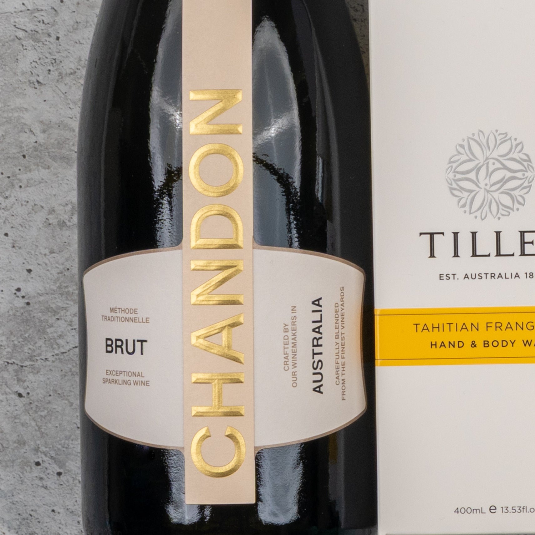 A Chandon Bubbles and Treats Hamper from The Hamper Boutique Co sits on a gray stone surface, featuring Chandon Brut sparkling wine alongside Tilley Tahitian Frangipani hand and body wash.