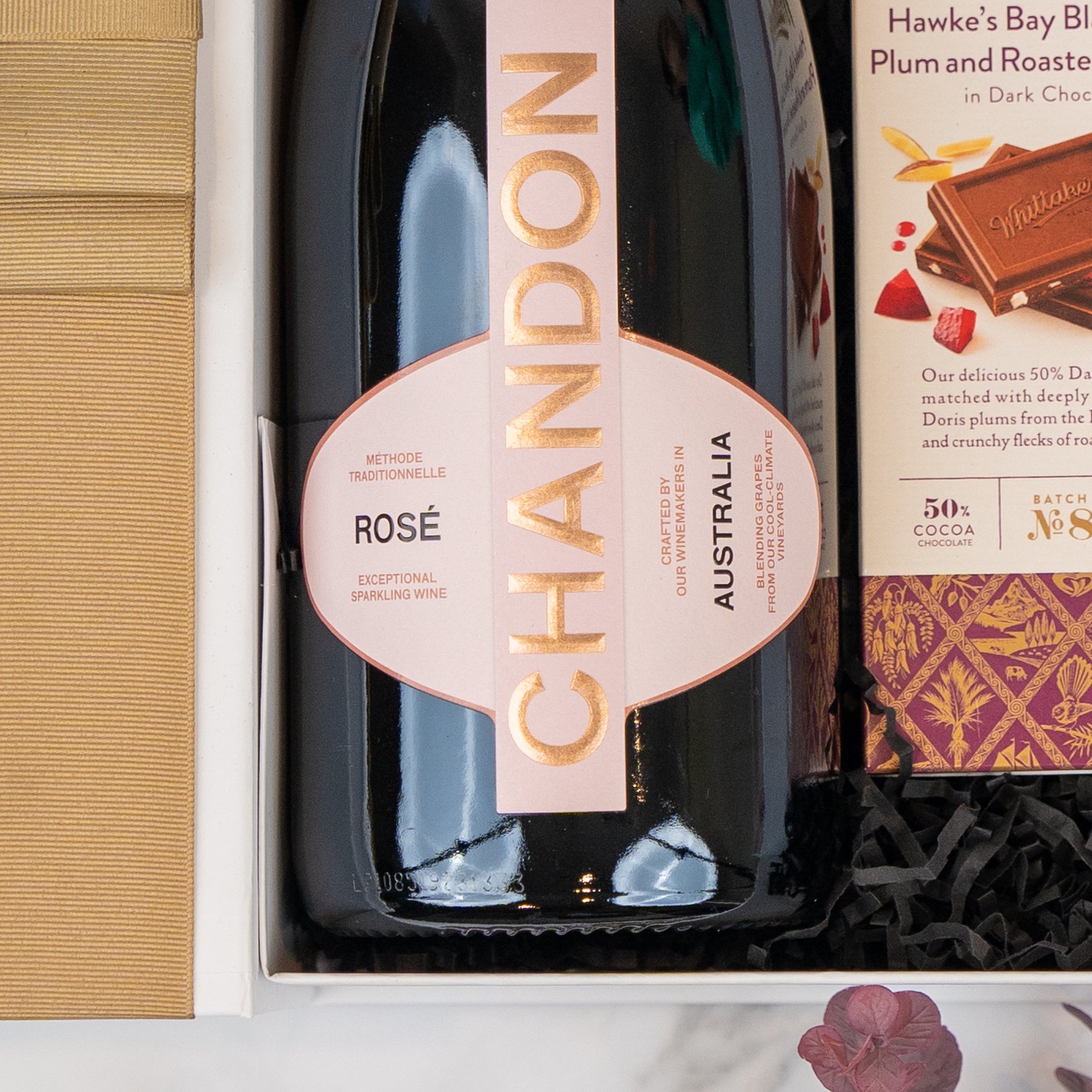 The Hamper Boutique Co Chandon Rosé Celebration Hamper includes an Australian Chandon Rosé sparkling wine in a gift box, paired with a chocolate bar in gold packaging and black shredded paper.