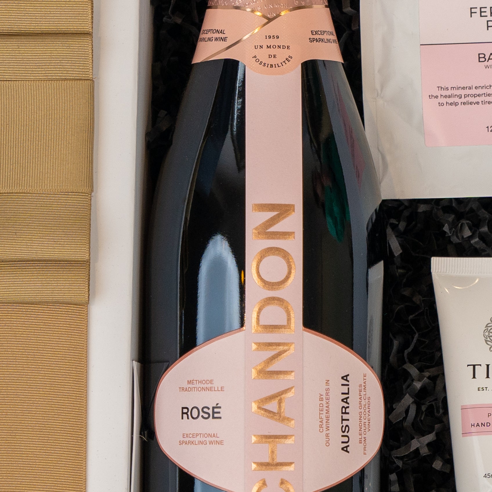 A close-up of The Hamper Boutique Co's Chandon Rosé Indulgence Hamper, featuring a bottle of Chandon Rosé with a pink and gold label nestled in shredded black paper, with other luxury gift items partially visible nearby.