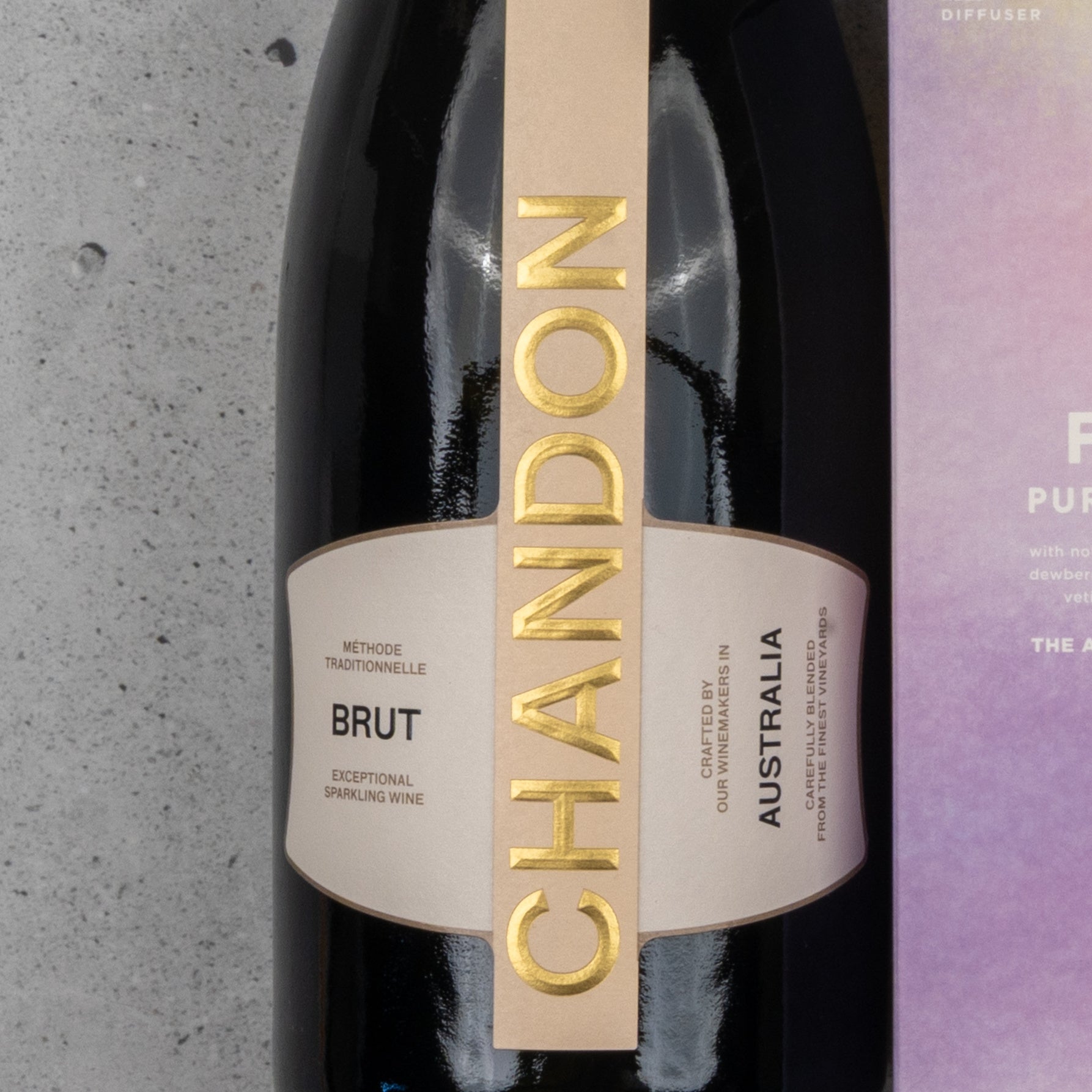 A close-up of The Hamper Boutique Co's Chandon Deluxe Celebration Hamper, featuring a bottle of sparkling wine with a gold label beside a purple box on a gray textured surface.