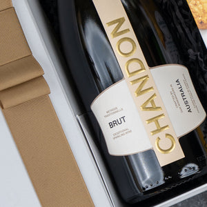 A close-up of The Hamper Boutique Co's Chandon Gourmet Sparkling Gift Hamper features a bottle of sparkling wine in a gift box beside a light brown ribbon and textured packaging material.