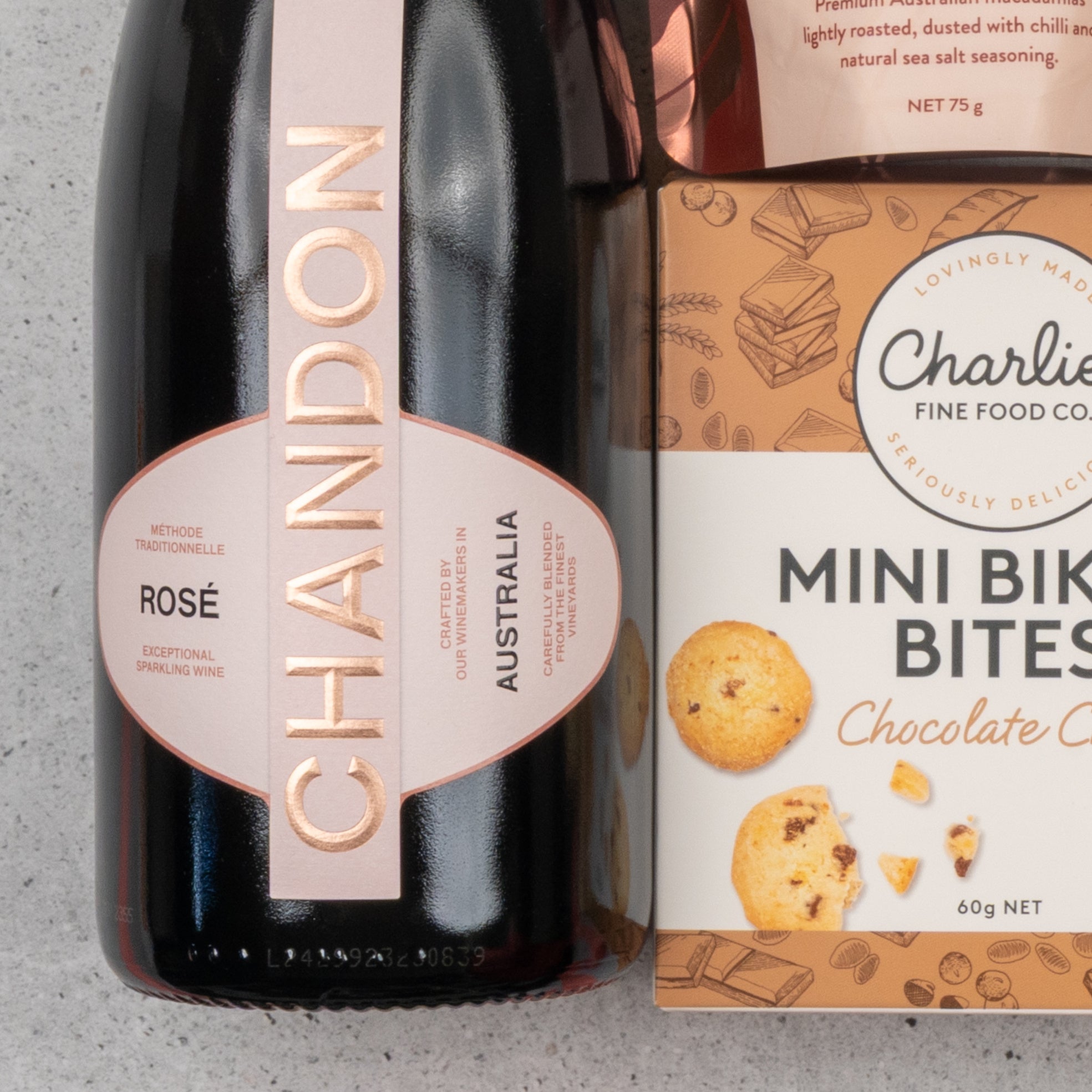 A Chandon Rosé Deluxe Celebration Hamper by The Hamper Boutique Co sits on a light-colored countertop, showcasing sparkling wine and Charlies Mini Bikkie Bites Chocolate Chip cookies.