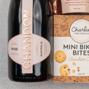 A Chandon Rosé Deluxe Celebration Hamper by The Hamper Boutique Co sits on a light-colored countertop, showcasing sparkling wine and Charlies Mini Bikkie Bites Chocolate Chip cookies.