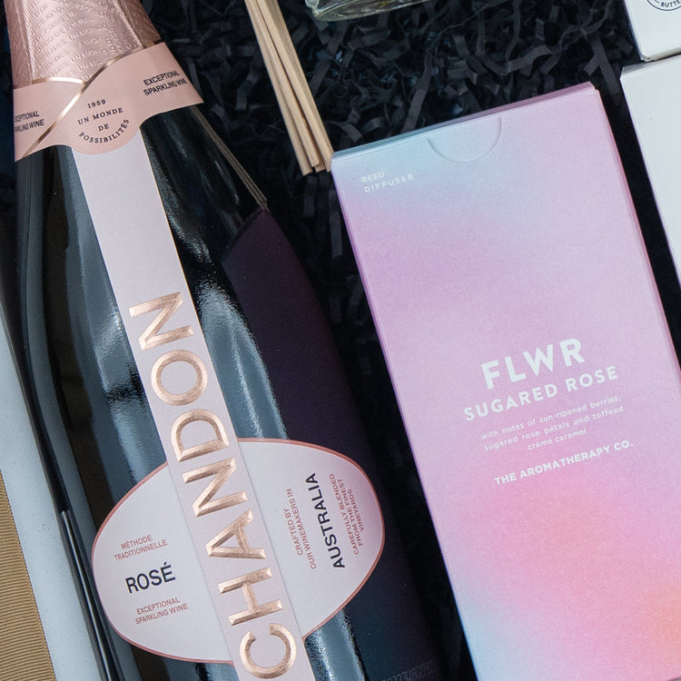 The Hamper Boutique Co's Chandon Connoisseur's Selection Hamper features a bottle of sparkling wine and a FLWR Sugared Rose Reed Diffuser in a pink box, arranged on black shredded paper inside an elegant gift box.