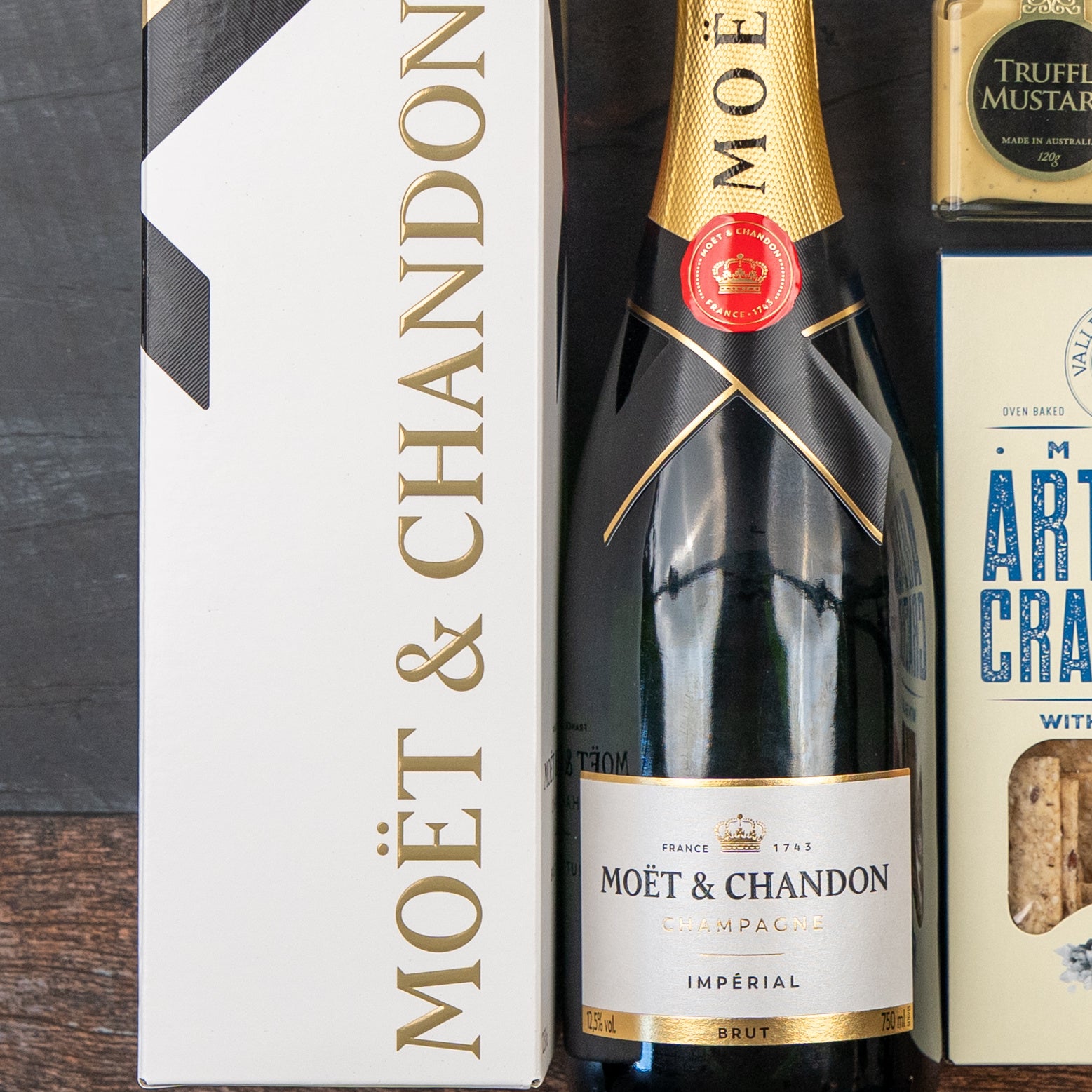 The Hamper Boutique Co's Moët & Chandon Celebration Hamper, featuring a boxed bottle of champagne, artisan crackers, and truffle mustard, is elegantly arranged on a dark wooden surface.