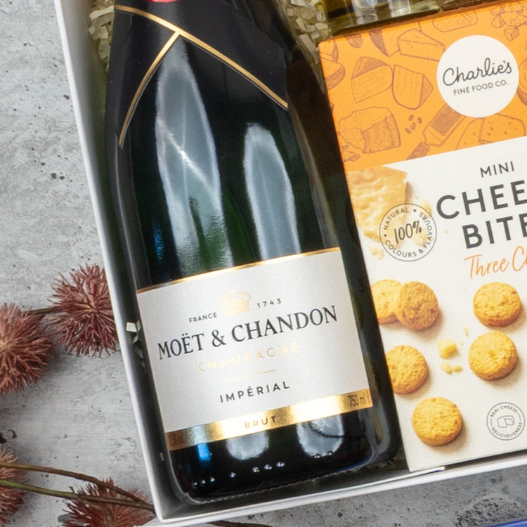 The Hamper Boutique Co’s Moët & Chandon Deluxe Celebration Hamper features a bottle of Moët & Chandon Champagne and Charlies mini cheese bites, beautifully presented on a light surface with dried flowers nearby.