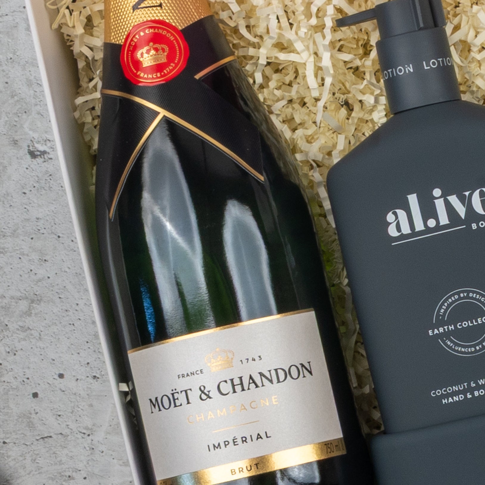 A Champagne Wellness Escape hamper by The Hamper Boutique Co, featuring a bottle of champagne next to a black al.ive pump bottle on shredded paper packing material.