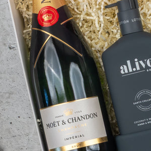 A Champagne Wellness Escape hamper by The Hamper Boutique Co, featuring a bottle of champagne next to a black al.ive pump bottle on shredded paper packing material.