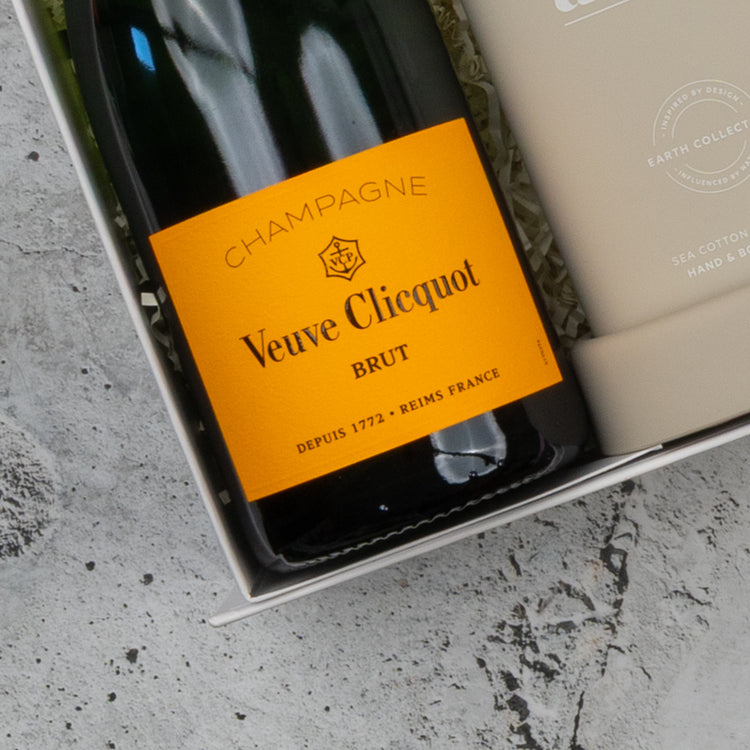 The Hamper Boutique Co's Veuve Clicquot Premium Celebration Hamper, displayed in its gift box beside a beige item with white text, rests on a light gray textured surface.