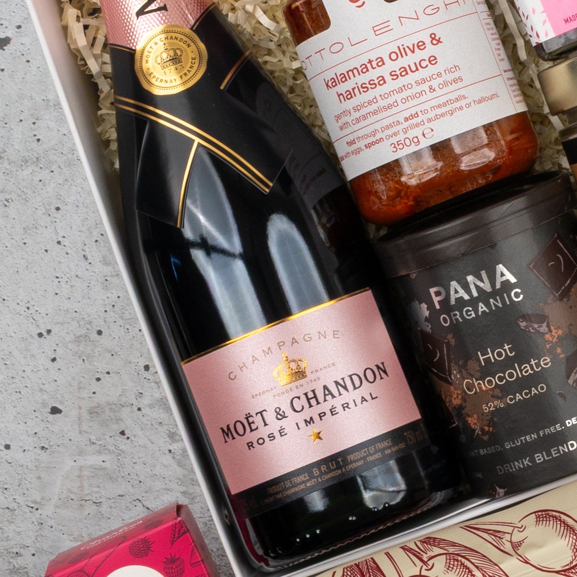 A close-up of The Hamper Boutique Co’s Moët & Chandon Rosé Grand Celebration Hamper, featuring champagne, kalamata olive & harissa sauce, and Pana Organic hot chocolate nestled on shredded paper.
