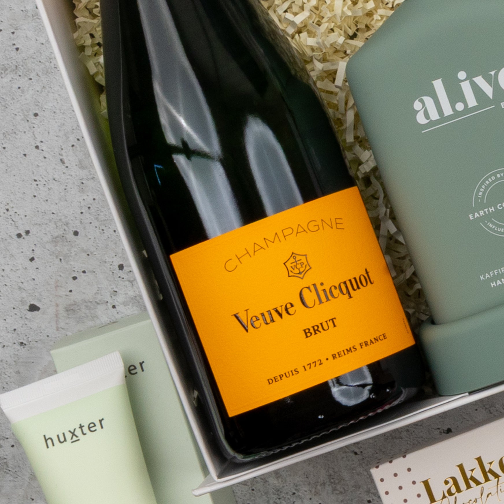 Close-up of The Hamper Boutique Co's Veuve Clicquot Grand Celebration Hamper, featuring a bottle of Veuve Clicquot Brut Champagne, a green Huxter tube, an al.ive green bottle, and shredded paper in the gift box.