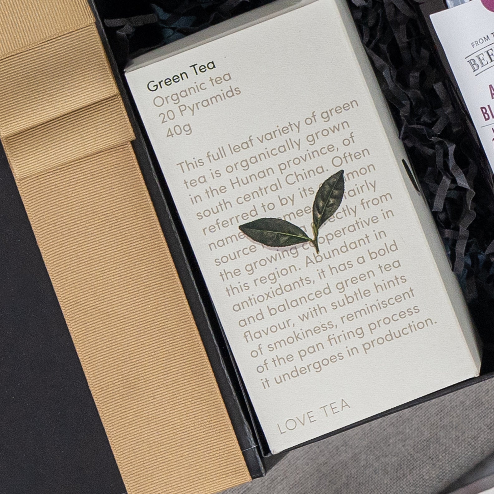 The Self-Care and Relaxation Hamper by The Hamper Boutique Co includes a gift box with organic green tea (20 pyramids, 40g), featuring details on origin, flavor, and antioxidants. A small decorative leaf adorns the elegant packaging.