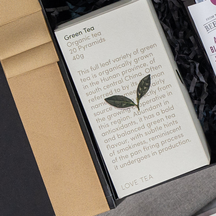 The Self-Care and Relaxation Hamper by The Hamper Boutique Co includes a gift box with organic green tea (20 pyramids, 40g), featuring details on origin, flavor, and antioxidants. A small decorative leaf adorns the elegant packaging.