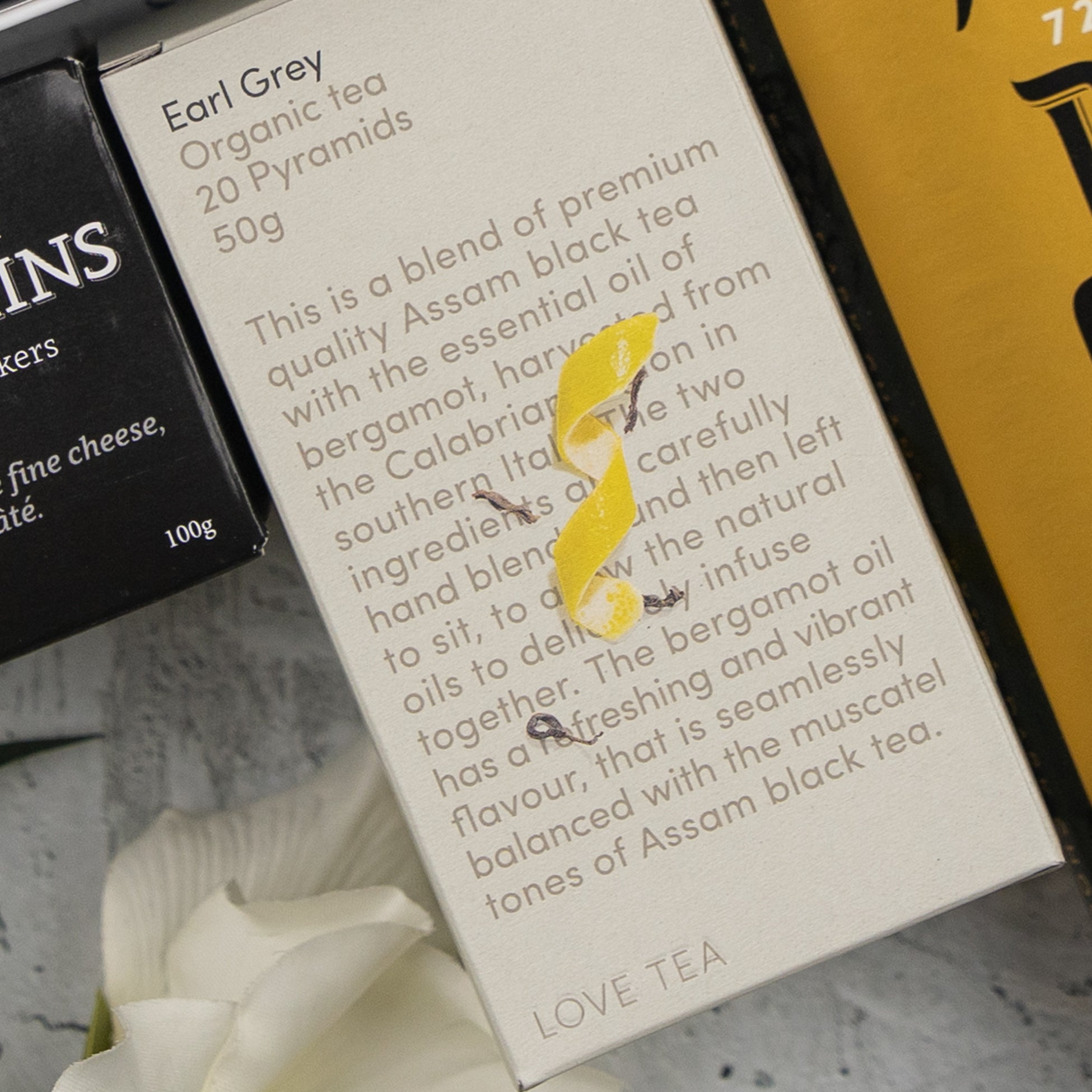 A box of The Hamper Boutique Co Black Tea Haven Hamper sits among other products, featuring 20 pyramid Assam black tea bags with bergamot and yellow string detail on the packaging.