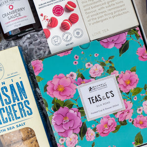 A top-down view of The Hamper Boutique Co's Sweet Indulgence Tea Hamper, featuring cranberry sauce, red macarons, crackers, and a floral-patterned "Teas & Cs Silk Road" tea gift box neatly arranged together.