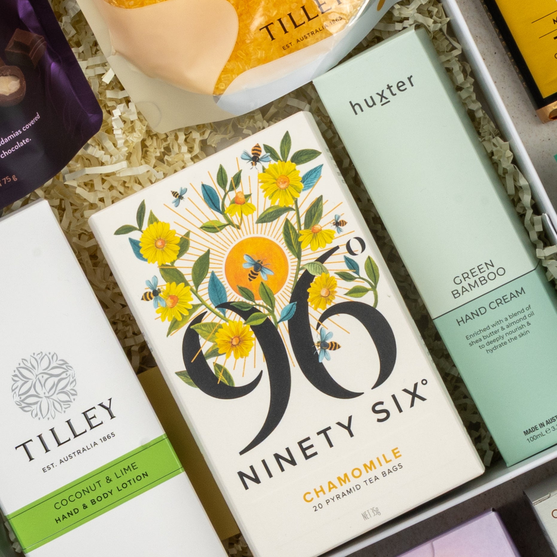 A close-up of The Hamper Boutique Co's Wellness Home Entertaining Hamper featuring chamomile tea, coconut & lime lotion, and green bamboo hand cream in yellow floral packaging on white shredded paper.