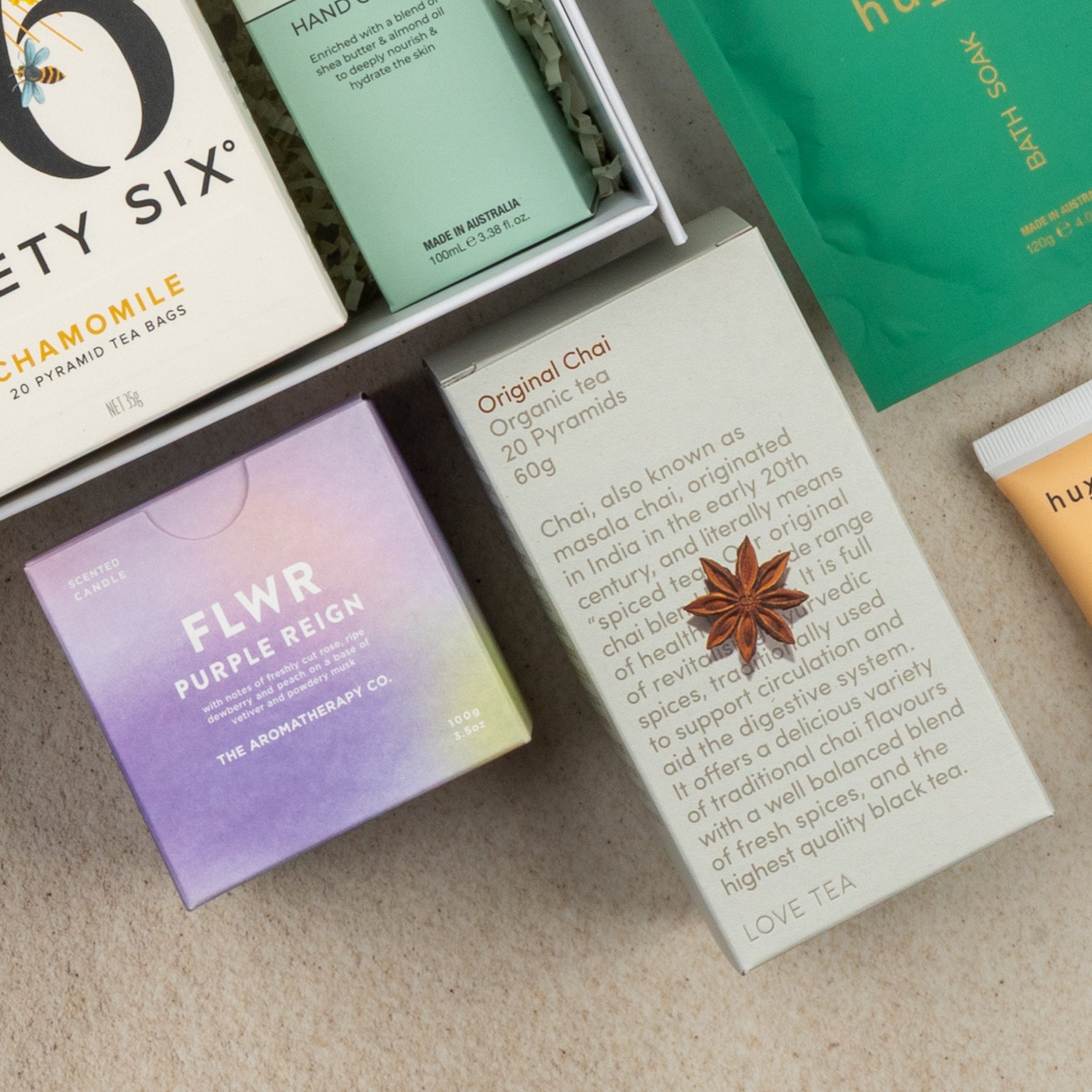 A flat lay of The Hamper Boutique Co's Wellness Home Entertaining Hamper, featuring chamomile tea, a FLWR Purple Reign box, Original Chai with anise star, and green bath soak packaging on a beige surface.
