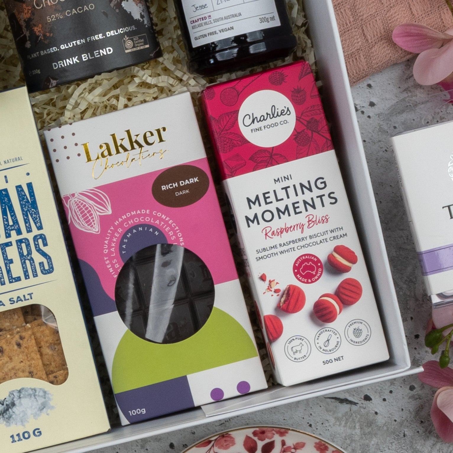The Hamper Boutique Co Premium Tea Experience Hamper features crackers, a Lakker Chocolates rich dark chocolate bar, and Charlies Mini Melting Moments Raspberry Bliss biscuits, beautifully arranged with shredded paper and pink flowers.