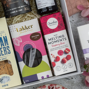 The Hamper Boutique Co Premium Tea Experience Hamper features crackers, a Lakker Chocolates rich dark chocolate bar, and Charlies Mini Melting Moments Raspberry Bliss biscuits, beautifully arranged with shredded paper and pink flowers.