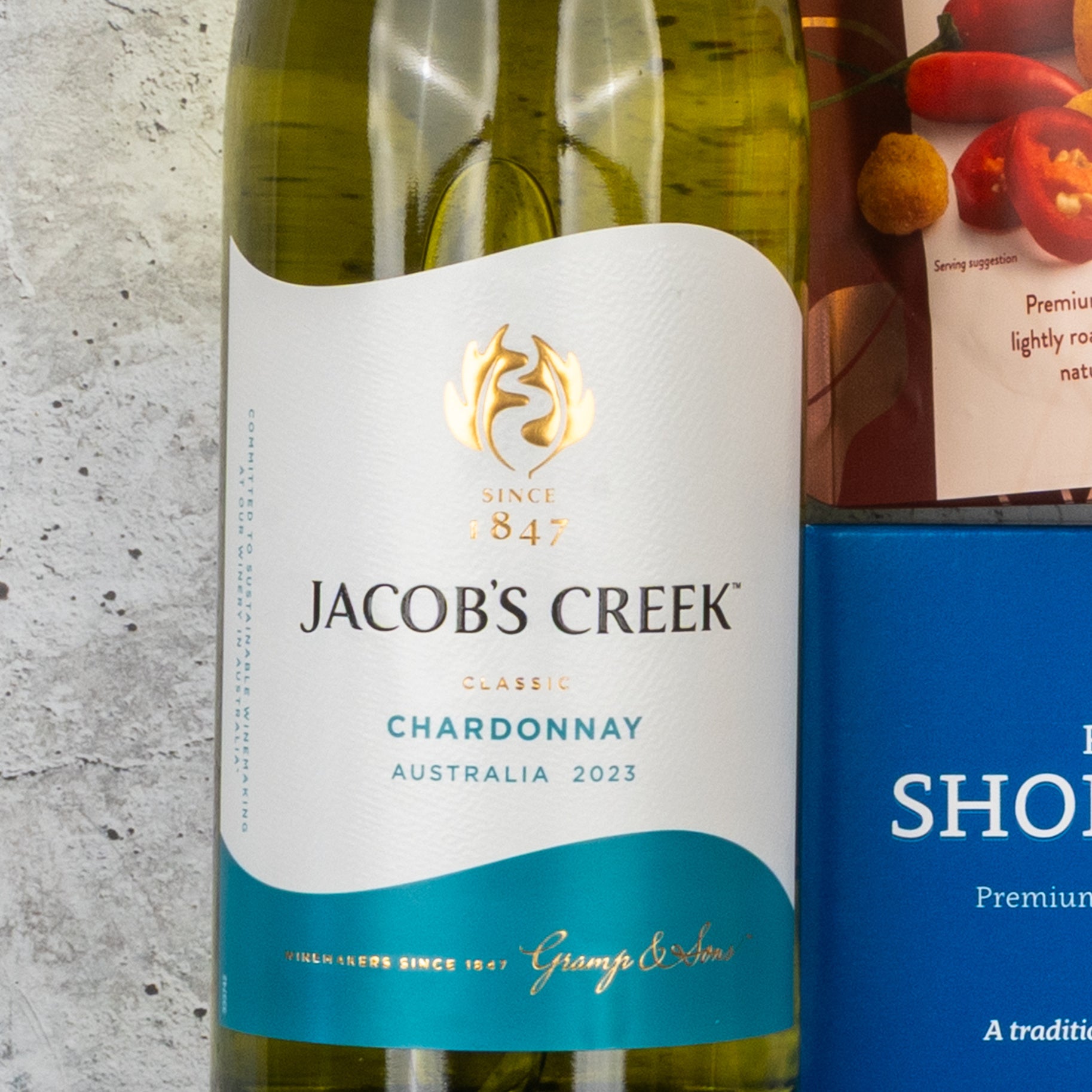 A Gourmet Getaway Gift Hamper by The Hamper Boutique Co is placed on a light stone surface, beside a blue rectangular item and part of a red-labeled food package.