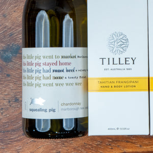 A Chardonnay and Self-Care Essentials hamper from The Hamper Boutique Co sits on a wooden surface, featuring a lively-labeled bottle of Chardonnay alongside Tilley Tahitian Frangipani hand and body lotion.