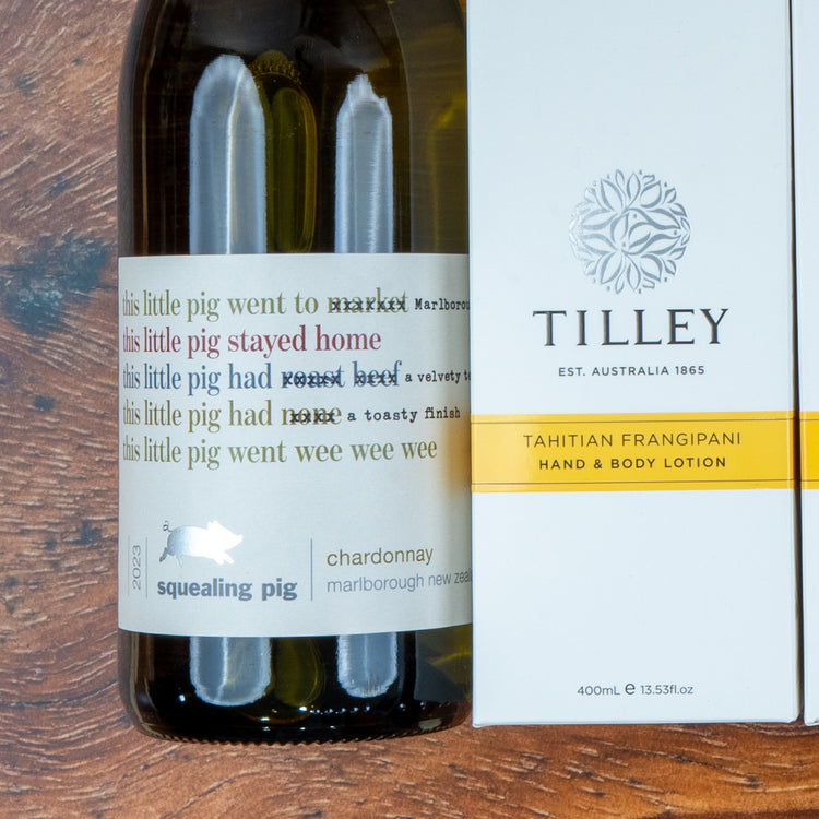 A Chardonnay and Self-Care Essentials hamper from The Hamper Boutique Co sits on a wooden surface, featuring a lively-labeled bottle of Chardonnay alongside Tilley Tahitian Frangipani hand and body lotion.