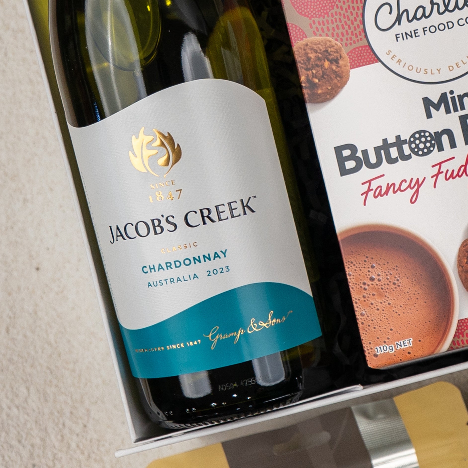 A Gourmet Indulgence Hamper from The Hamper Boutique Co features a white and teal wine bottle beside a box of chocolate mint button cookies, with the round treats partially visible.