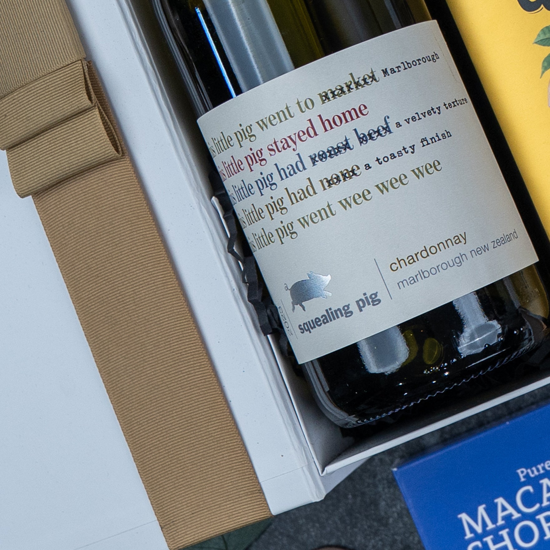 A Gourmet Experience Hamper by The Hamper Boutique Co features a bottle of chardonnay wine in a gift box, shown alongside a wrapped present and part of a blue MACAR box. The wine label displays playful, colorful text.