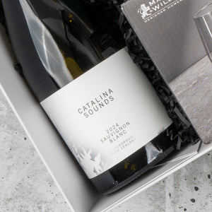 A bottle from The Hamper Boutique Co’s Chardonnay and Relaxation Gift Hamper lies partially tilted in a gift box with crinkled black paper and the corner of a nearby booklet visible.
