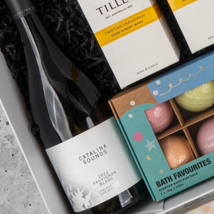A close-up of The Hamper Boutique Co's Ultimate Self-Care and Chardonnay Hamper, featuring a bottle of Chardonnay, Tillie hand and body wash, and Bath Favourites bath bombs arranged on shredded black paper.