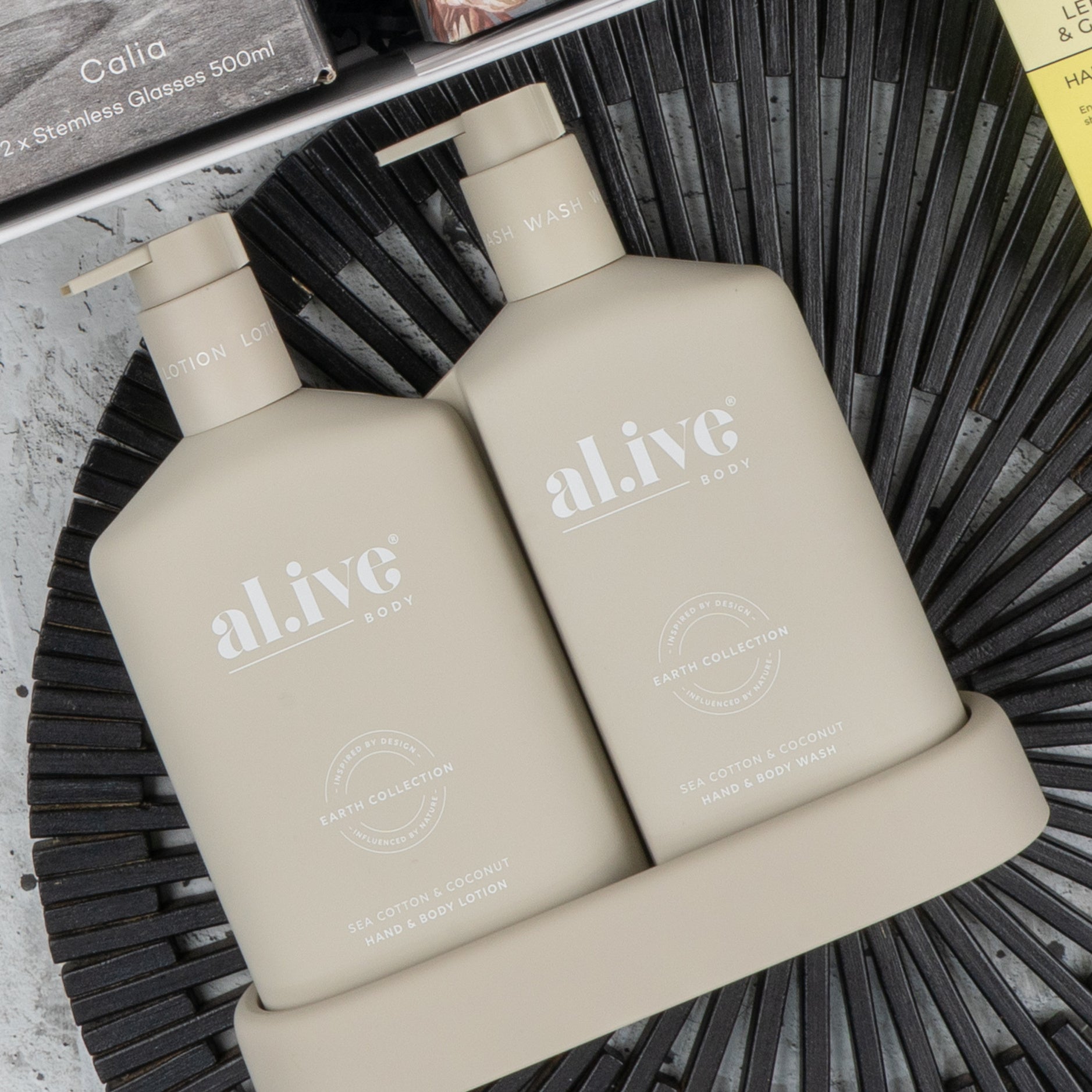 Two beige bottles of The Hamper Boutique Co’s Elegance and Self-Care Gift Hamper, featuring lotion and body wash in “Sea Cotton & Coconut” from the Earth Collection, sit on a matching tray atop a black textured surface.
