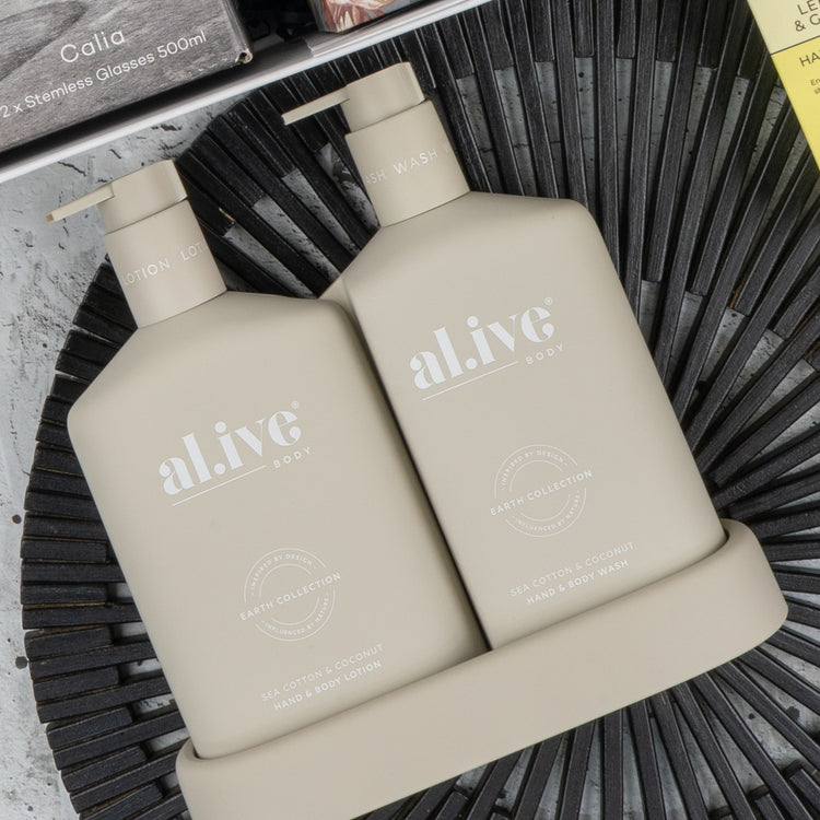 Two beige bottles of The Hamper Boutique Co’s Elegance and Self-Care Gift Hamper, featuring lotion and body wash in “Sea Cotton & Coconut” from the Earth Collection, sit on a matching tray atop a black textured surface.