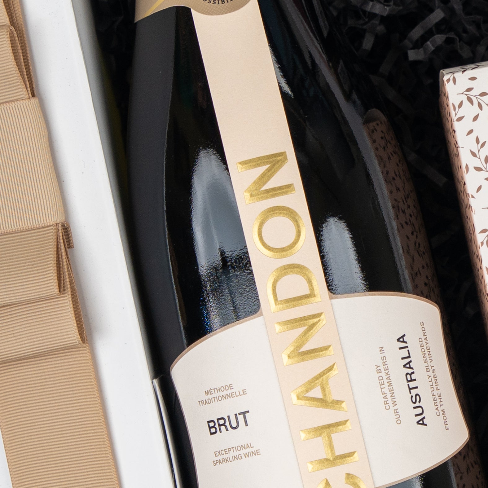 A close-up of The Hamper Boutique Co's Chandon Sparkling Gourmet Hamper, featuring a bottle of Chandon sparkling wine nestled in a gift box with a beige ribbon, and an elegant patterned box beside it.