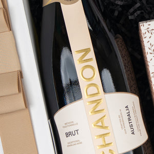 A close-up of The Hamper Boutique Co's Chandon Sparkling Gourmet Hamper, featuring a bottle of Chandon sparkling wine nestled in a gift box with a beige ribbon, and an elegant patterned box beside it.