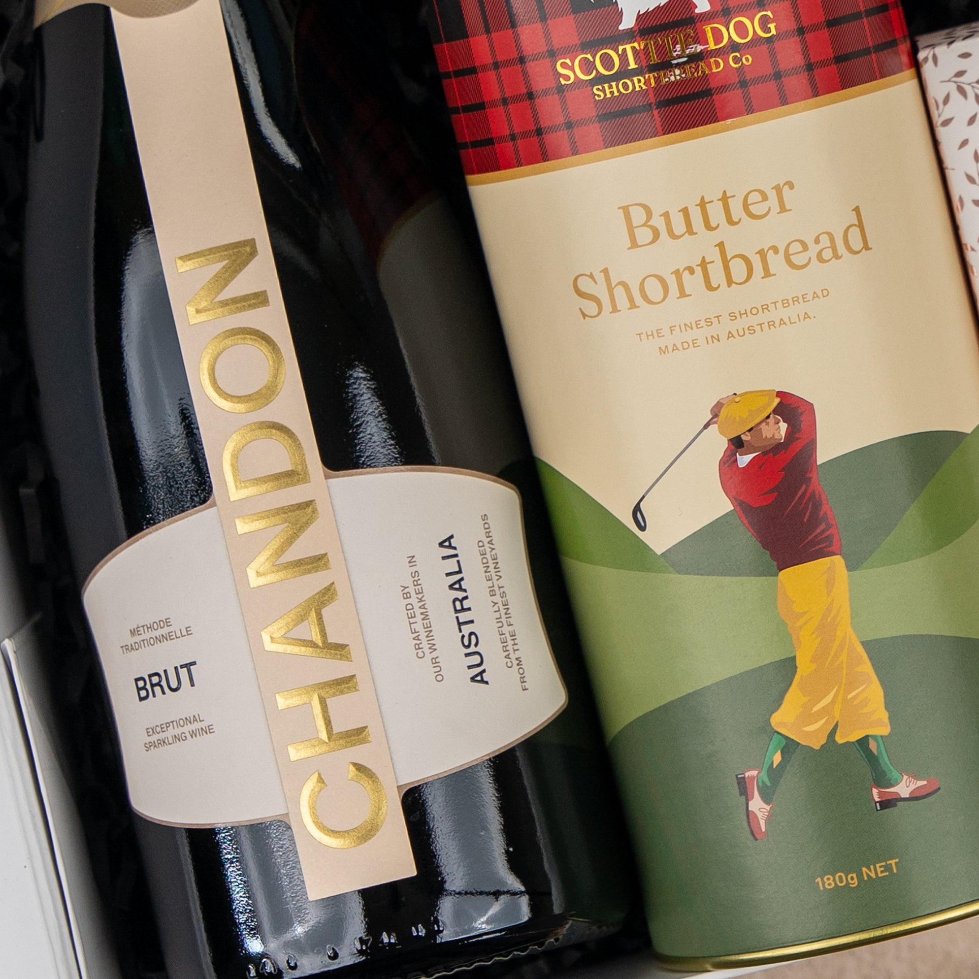 A bottle from The Hamper Boutique Co's Sparkling Comfort & Joy sits beside a Scottie Dog Butter Shortbread tin, which is decorated with an illustration of a golfer swinging a club.