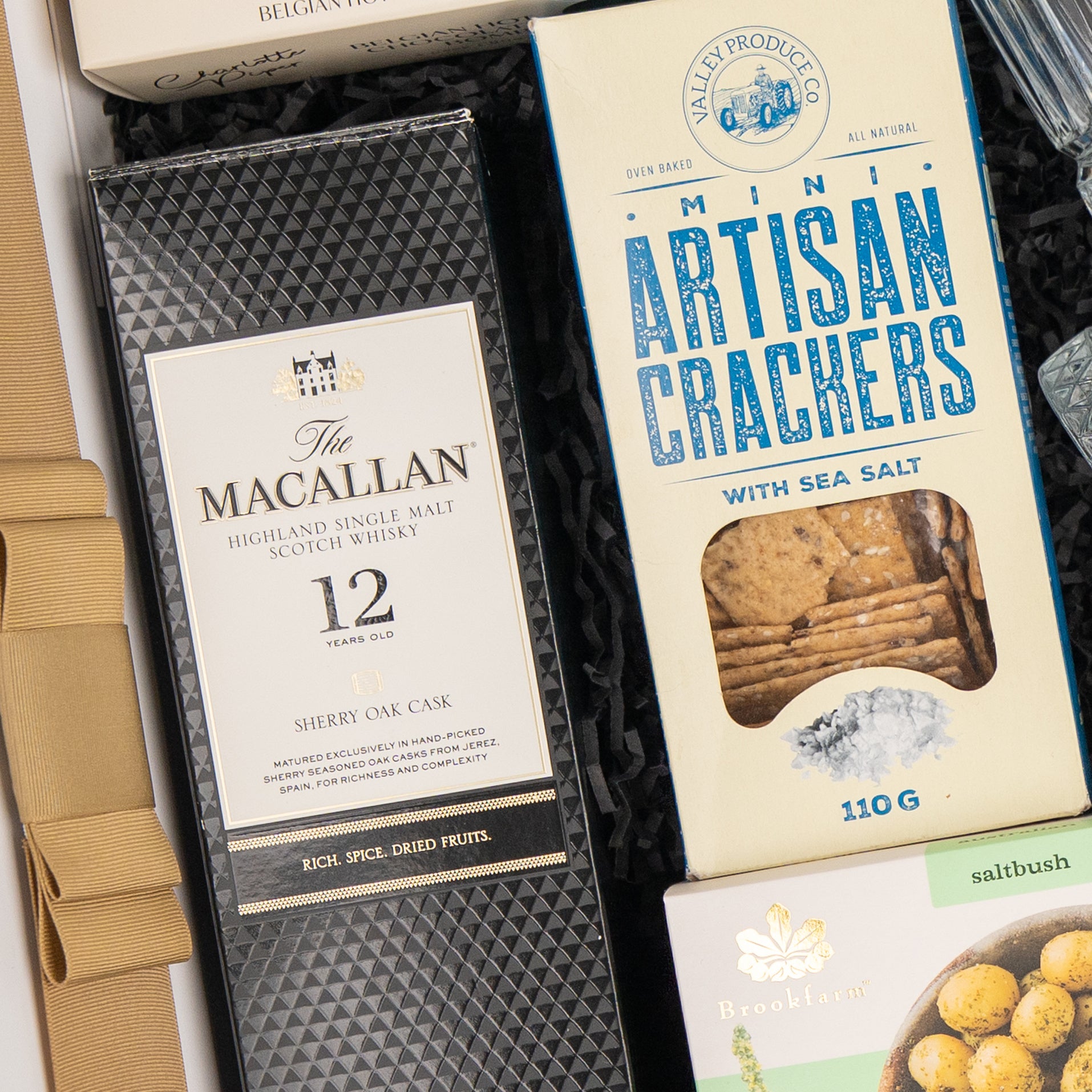 The Hamper Boutique Co's Whisky Celebration Extravaganza features The Macallan 12-Year-Old Scotch, mini sea salt artisan crackers, and Brookfarm nuts, all beautifully arranged on black shredded paper.