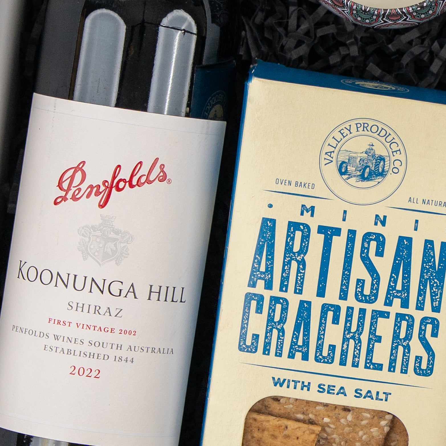 A Wine, Candle and Board Hamper from The Hamper Boutique Co is displayed next to a blue and cream box of Valley Produce Co. Mini Artisan Crackers with Sea Salt.