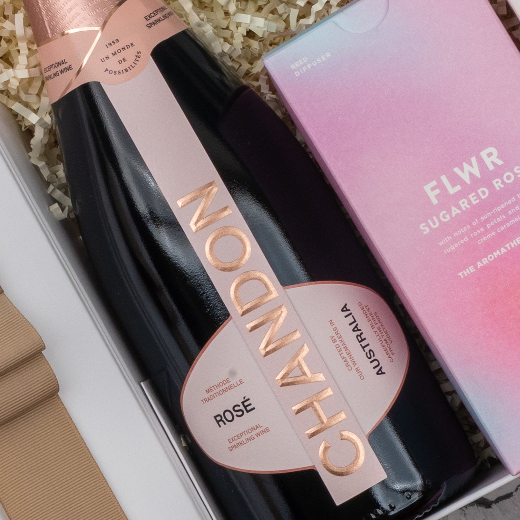 A Chandon Rosé Pamper Hamper by The Hamper Boutique Co rests on shredded packing, with a bottle of sparkling wine beside a pink FLWR Sugared Rose box.