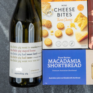 A White Wine Gourmet Hamper by The Hamper Boutique Co sits on a dark marble surface, featuring chardonnay, Mini Cheese Bites, and Pure Butter Macadamia Shortbread.