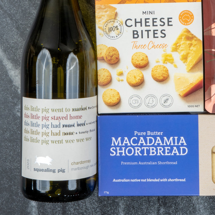 A White Wine Gourmet Hamper by The Hamper Boutique Co sits on a dark marble surface, featuring chardonnay, Mini Cheese Bites, and Pure Butter Macadamia Shortbread.