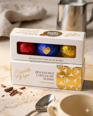 Two boxes of Charlotte Piper Belgian Chocolate Bombs Mixed, each with three foil-wrapped chocolates in red, blue, and gold, are displayed on a marble surface near a cup, chocolate shavings, and a metal jug.
