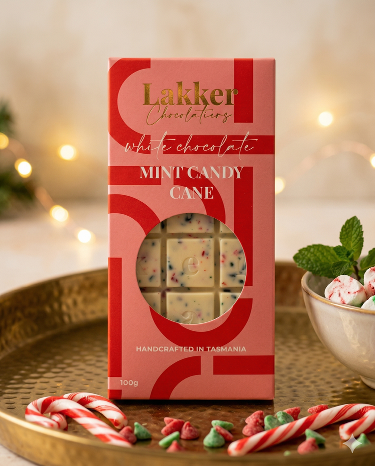 A box of Lakker Chocolatiers Mint Candy Cane is displayed on a gold tray with candy canes, mint candies, and festive lights with greenery in the background.