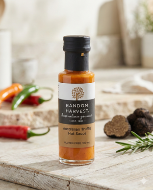 A bottle of Random Harvest Australian Truffle Hot Sauce rests on a rustic kitchen counter, surrounded by fresh red chilies, green herbs, and whole black truffles, with kitchenware softly blurred in the background.