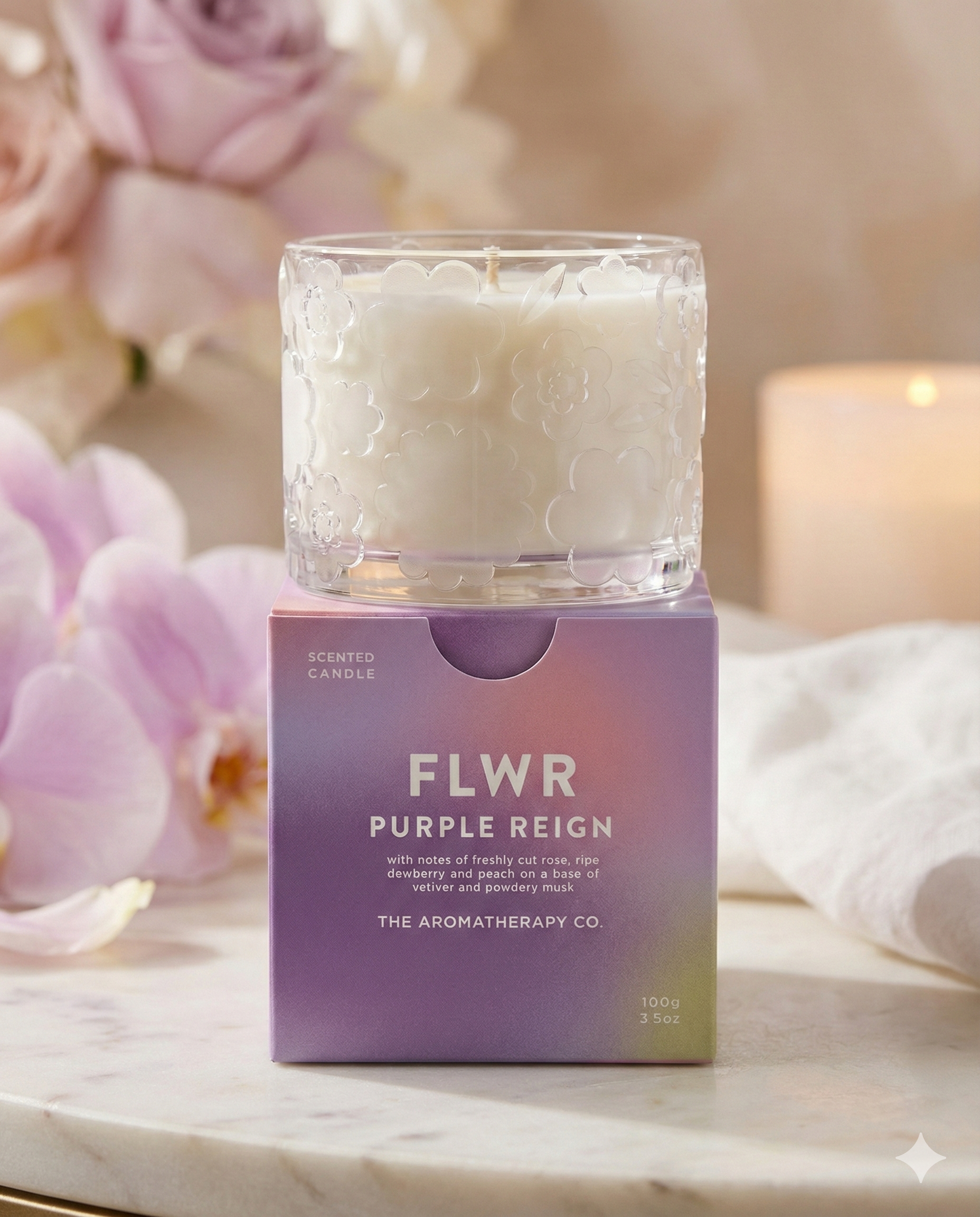 The Flwr Purple Reign Candle by The Aromatherapy Company, in a glass jar with a floral design, sits on a purple ombre box surrounded by soft pink flowers and a softly blurred candle in the background.